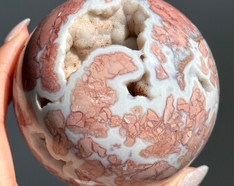 May include: A spherical, polished gemstone with a geode cavity. The orb features a mix of pink, white, and tan colors, with a cracked, mosaic-like pattern across its surface. The geode interior is a light tan color.