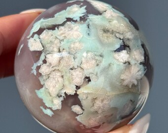 May include: A polished, spherical gemstone with a mix of colors. The base color is a soft lavender, overlaid with patches of pale green and creamy white, creating a unique, marbled appearance. The sphere is held by a person.