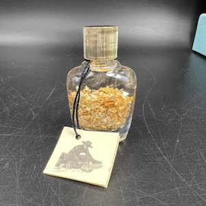 May include: A clear glass bottle filled with gold flakes, with a gold-colored cap and a black string attached to a tag. The tag has a black and white illustration of a person panning for gold.