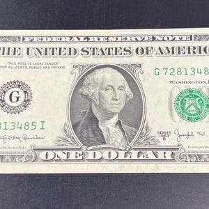 May include: A one dollar bill featuring a portrait of George Washington. The bill is green and white with the text "THE UNITED STATES OF AMERICA" and "ONE DOLLAR" printed on it. The serial number is G72813485 I.