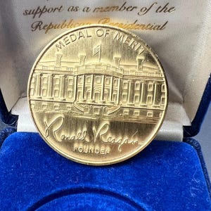 May include: A gold-colored Medal of Merit, featuring an image of a building and the text "MEDAL OF MERIT" and "Ronald Reagan FOUNDER". The medal is displayed in a blue velvet box with gold lettering.