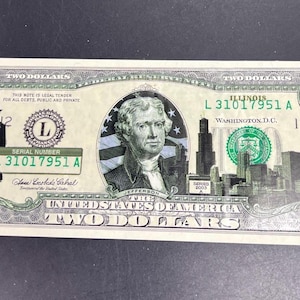 May include: A United States two-dollar bill. The bill features a portrait of Thomas Jefferson, the text "TWO DOLLARS", and a silhouette of a man. The serial number is L31017951A. The bill also has a depiction of a city skyline.
