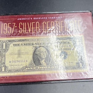 May include: A 1957 Silver Certificate one-dollar bill in a protective case. The bill features a portrait of George Washington and the text "The United States of America." The case is red with the text "America's Obsolete Currency" and "1957 Silver Certificate."