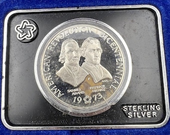 May include: A silver coin commemorating the American Revolution Bicentennial, featuring portraits of Samuel Adams and Patrick Henry. The coin is set in a black rectangular frame with the words "STERLING SILVER" and the year 1973.