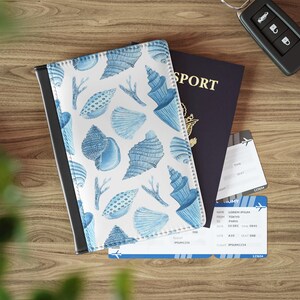 May include: A passport cover with a white background and blue seashell pattern, next to a dark blue passport, plane tickets, and a car key. The passport cover has a black border. The passport says "SPORT" in gold letters.