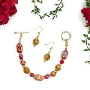 May include: A gold-toned jewelry set featuring a bracelet and two earrings. The bracelet has a toggle clasp and is adorned with a mix of amber, red, and gold beads. The earrings have a similar design with amber-colored beads.