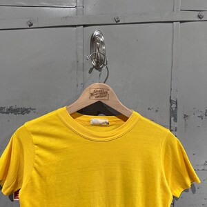 May include: A bright yellow short-sleeved t-shirt hanging on a wooden hanger. The hanger has the text "Great Northern Vintage" printed on it. The shirt is made of a soft material.