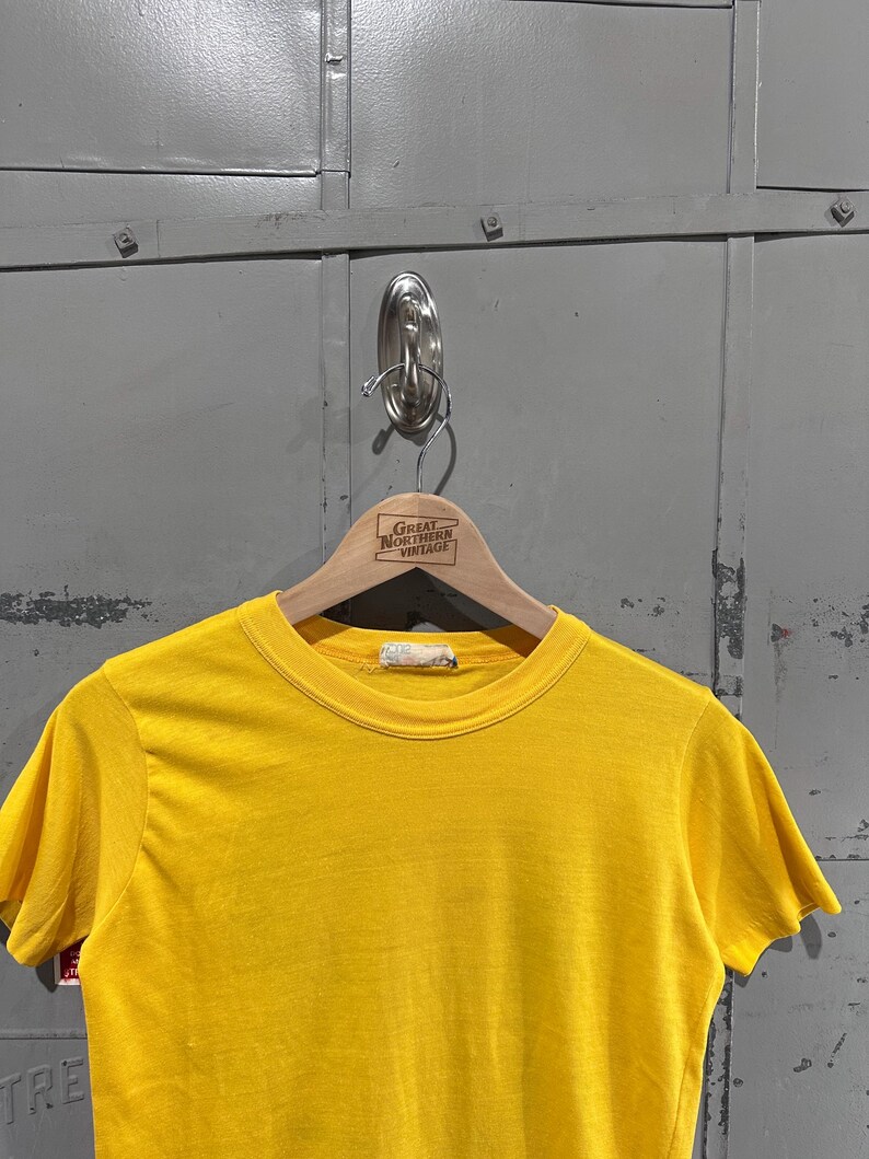 May include: A bright yellow short-sleeved t-shirt hanging on a wooden hanger. The hanger has the text "Great Northern Vintage" printed on it. The shirt is made of a soft material.