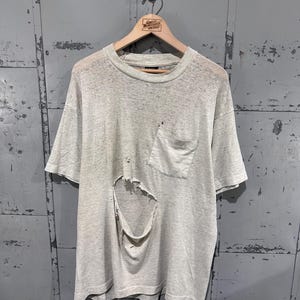 May include: A light gray heathered short-sleeve t-shirt featuring a crew neckline and a single chest pocket on the left side. The garment displays significant wear, including a large, prominent tear in the center of the front torso area.