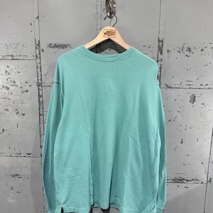 Könnte beinhalten: Ein langärmeliges Shirt mit Rundhalsausschnitt in einem einfarbigen hellen Mintgrün. Das Shirt verfügt über einen gerippten Halsausschnitt, lange Ärmel mit gerippten Bündchen und kleine Seitenschlitze am unteren Saum. Der Stoff ist glatt.