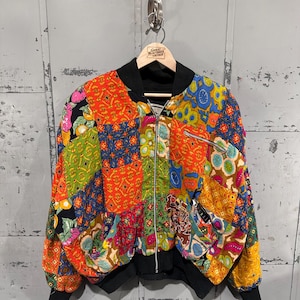 May include: A colorful patchwork bomber jacket with a black collar, cuffs, and hem. The jacket features a variety of patterned fabric squares in shades of orange, blue, green, and yellow. It has a front zipper and a pocket on the left sleeve.