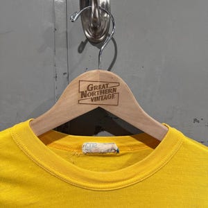 May include: A bright yellow t-shirt hangs on a wooden hanger. The hanger has the text "GREAT NORTHERN VINTAGE" printed on it. The shirt is hanging on a metal hook attached to a gray wall.