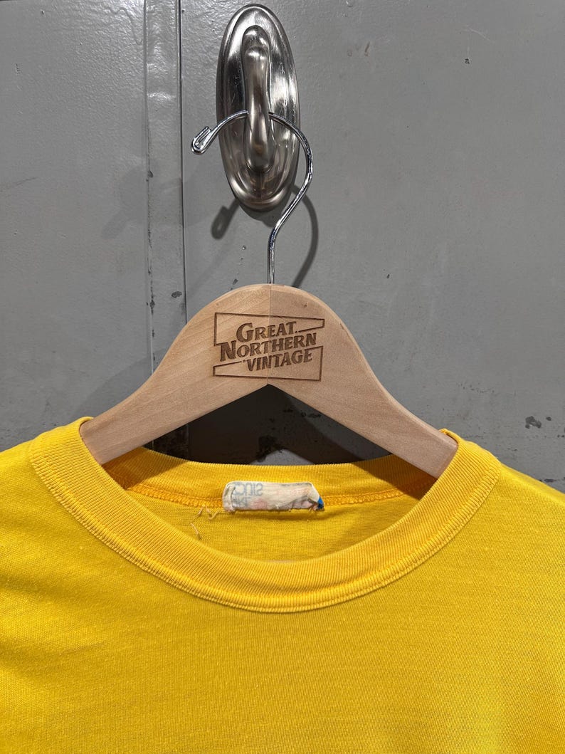 May include: A bright yellow t-shirt hangs on a wooden hanger. The hanger has the text "GREAT NORTHERN VINTAGE" printed on it. The shirt is hanging on a metal hook attached to a gray wall.