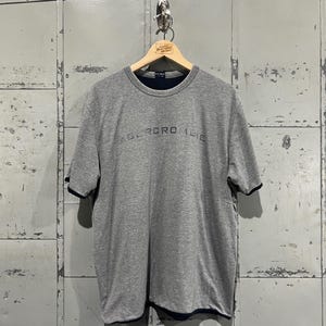 May include: A grey t-shirt with a navy blue trim around the neckline and sleeves. The shirt has the word "Abercrombie" printed in black on the front.
