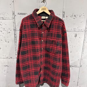 Può includere: Una camicia a quadri rossi e neri con bottoni, colletto e una tasca sul petto. La camicia è realizzata in un materiale soffice ed è appesa a una gruccia di legno.