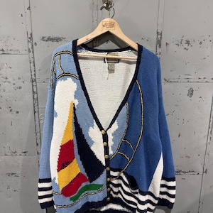 May include: A blue, white, and navy cardigan sweater with a sailboat design. The sweater has gold buttons and a V-neck. The sleeves and bottom hem have white and navy stripes. The sweater is hanging on a wooden hanger.