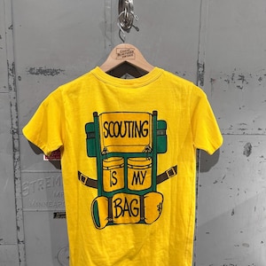 May include: A yellow t-shirt with a graphic of a green and yellow backpack. The backpack design includes the words "SCOUTING IS MY BAG" in black lettering. The shirt is hanging on a wooden hanger.