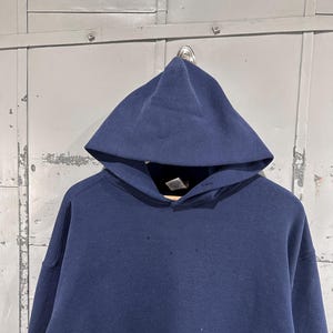 May include: A navy blue hooded sweatshirt with a drawstring neckline. The sweatshirt has a large hood and long sleeves. The fabric appears to be a thick, durable material. The garment is hanging on a metal hook against a gray background.