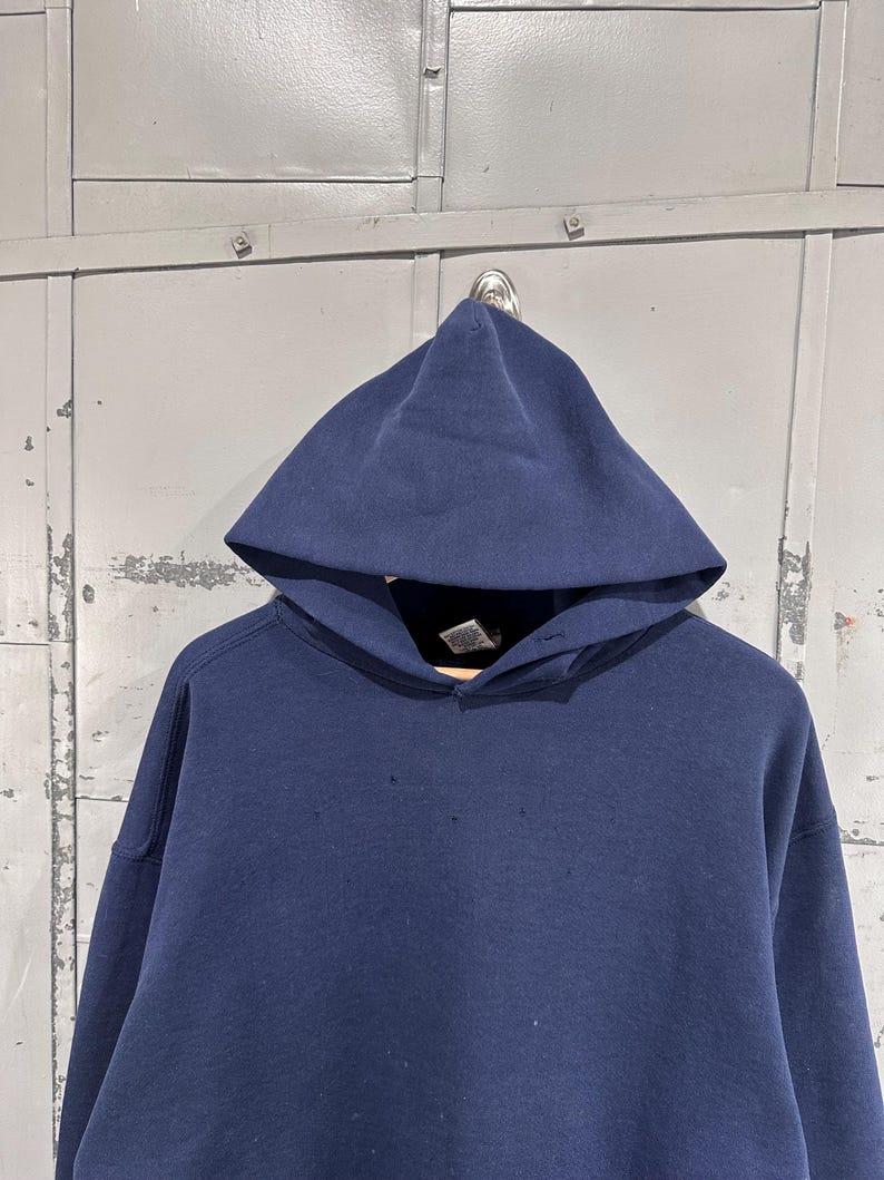May include: A navy blue hooded sweatshirt with a drawstring neckline. The sweatshirt has a large hood and long sleeves. The fabric appears to be a thick, durable material. The garment is hanging on a metal hook against a gray background.