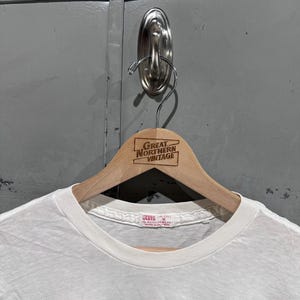 May include: A white t-shirt with the word "Regina" in blue lettering. The shirt is hanging on a wooden hanger with the text "Great Northern Vintage". The shirt has a crew neck and short sleeves.