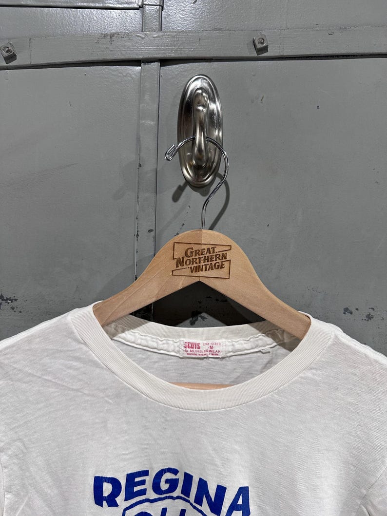 May include: A white t-shirt with the word "Regina" in blue lettering. The shirt is hanging on a wooden hanger with the text "Great Northern Vintage". The shirt has a crew neck and short sleeves.
