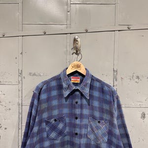 Pu&ograve; includere: Una camicia a maniche lunghe con bottoni e un motivo a quadri blu e azzurro. La camicia presenta un colletto a punta, due tasche sul petto con bottoni neri e un'abbottonatura centrale. Un'etichetta &egrave; visibile all'interno del colletto.