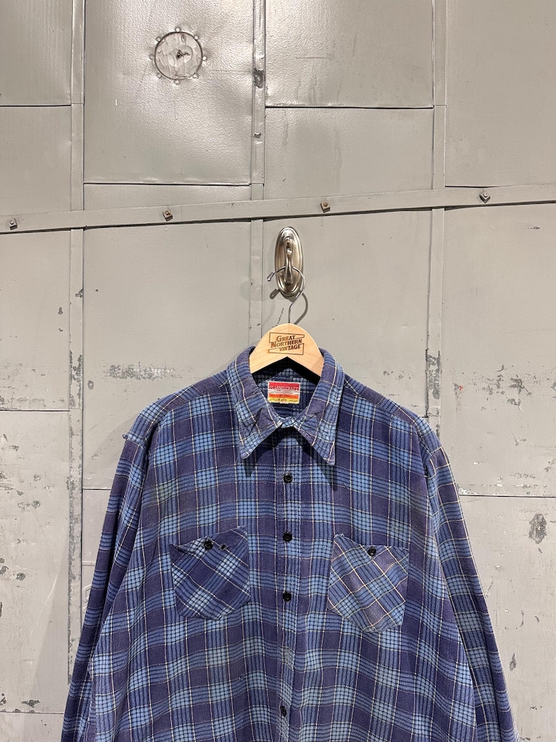 Pu&ograve; includere: Una camicia a maniche lunghe con bottoni e un motivo a quadri blu e azzurro. La camicia presenta un colletto a punta, due tasche sul petto con bottoni neri e un'abbottonatura centrale. Un'etichetta &egrave; visibile all'interno del colletto.