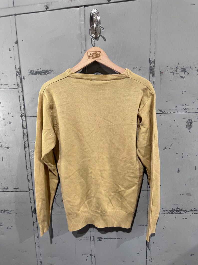 May include: A mustard yellow sweater with long sleeves, hanging on a wooden hanger. The hanger has the text "Great Northern Vintage" printed on it. The sweater is made of a knit material.