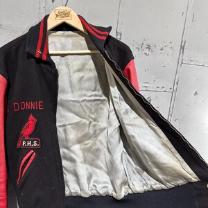 Op de afbeelding: Zwarte en rode vintage varsity jacket met de naam "Donnie" in het rood geborduurd. Voorzien van een kardinaalvogelpatch en de letters "P.H.S.". De jas heeft een zwarte geribbelde kraag, manchetten en tailleband, met rode leren mouwen en accenten.