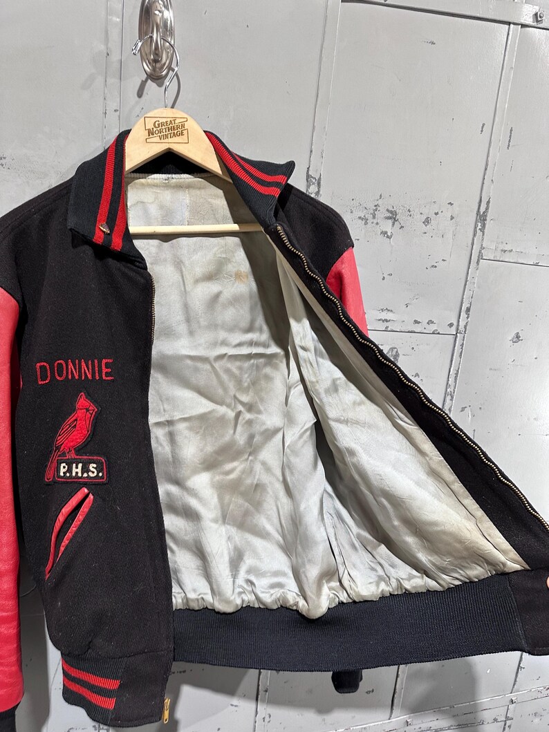 Op de afbeelding: Zwarte en rode vintage varsity jacket met de naam "Donnie" in het rood geborduurd. Voorzien van een kardinaalvogelpatch en de letters "P.H.S.". De jas heeft een zwarte geribbelde kraag, manchetten en tailleband, met rode leren mouwen en accenten.