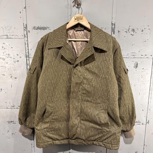 May include: An olive green vintage jacket with a vertical striped pattern. The jacket has a collared neckline, two front pockets, and ribbed cuffs. The jacket is hanging on a wooden hanger.