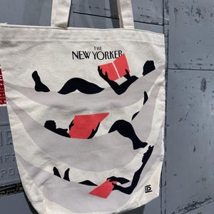 May include: Off-white canvas tote bag featuring the text "THE NEW YORKER" in black. The bag displays a graphic of figures reclining and reading red books. The tote has two handles and is made of a sturdy fabric.