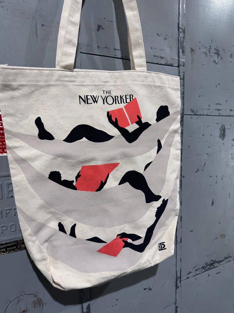 May include: Off-white canvas tote bag featuring the text "THE NEW YORKER" in black. The bag displays a graphic of figures reclining and reading red books. The tote has two handles and is made of a sturdy fabric.