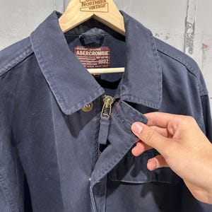 May include: Navy blue Abercrombie jacket with a zipper closure and a collared neckline. The jacket has a pocket on the front and a label that reads "Abercrombie 1892". The jacket is hanging on a wooden hanger.