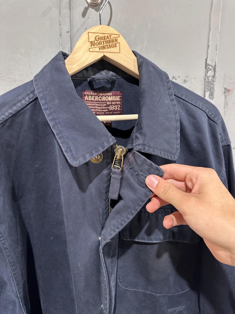 May include: Navy blue Abercrombie jacket with a zipper closure and a collared neckline. The jacket has a pocket on the front and a label that reads "Abercrombie 1892". The jacket is hanging on a wooden hanger.