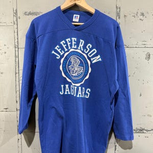 May include: A blue long-sleeve shirt with a v-neck collar. The shirt features a white circular emblem with the words "JEFFERSON" and "JAGUARS" in white. The emblem also includes the text "THOMAS JEFFERSON HIGH SCHOOL" and the year "1972".