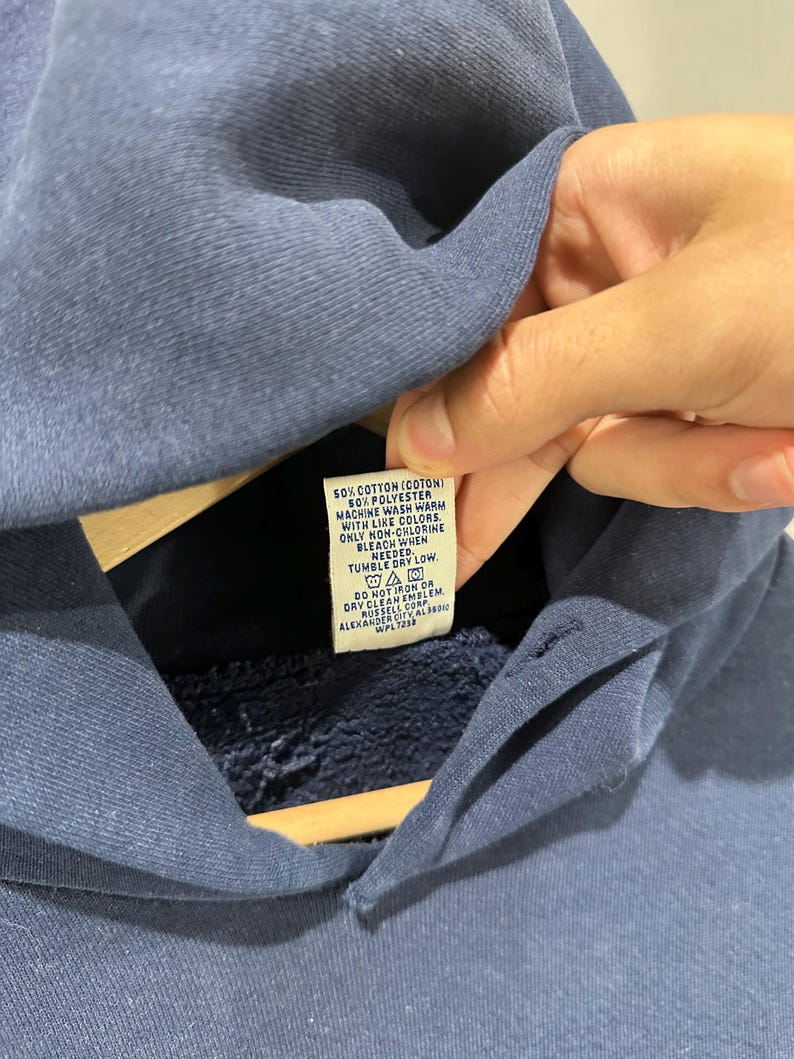 May include: A navy blue hoodie with a tag visible. The tag reads "50% Cotton (Coton) 50% Polyester Machine wash warm with like colors. Only non-chlorine bleach when needed. Tumble dry low."