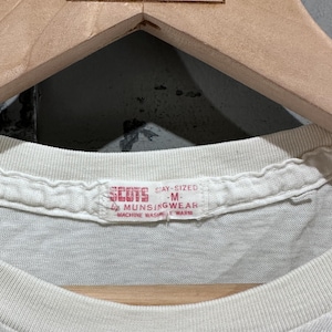 May include: Close-up of a vintage white t-shirt with a crew neck. The tag reads "SCOTS STAY-SIZED M by MUNSINGWEAR MACHINE WASH WARM". The shirt is hanging on a wooden hanger with the text "NORTHERN VINTAGE" at the top.