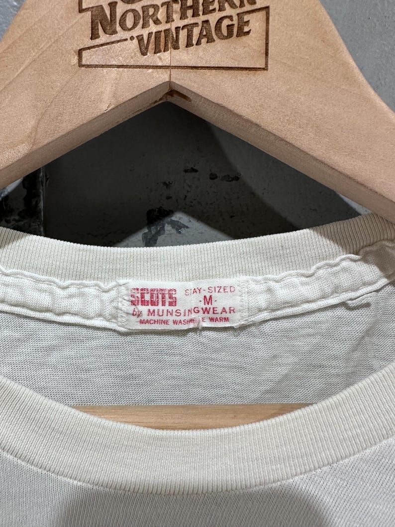 May include: Close-up of a vintage white t-shirt with a crew neck. The tag reads "SCOTS STAY-SIZED M by MUNSINGWEAR MACHINE WASH WARM". The shirt is hanging on a wooden hanger with the text "NORTHERN VINTAGE" at the top.