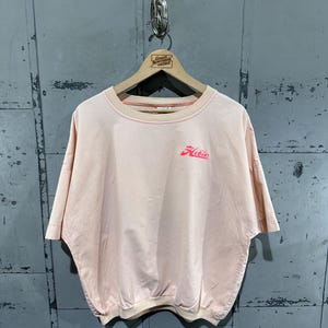 Könnte beinhalten: Hellrosa kurzärmeliges T-Shirt mit dem Wort "Hobie" in roter Schrift auf der Vorderseite. Das Shirt hat einen Rundhalsausschnitt und einen gerafften Saum. Das Shirt hängt an einem Holzbügel.