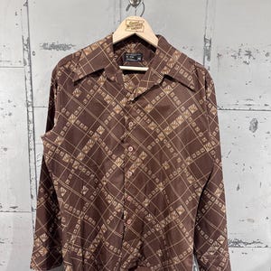 May include: A brown button-down shirt with a geometric pattern of tan lines and shapes. The shirt has a pointed collar and long sleeves. The shirt is hanging on a wooden hanger with the text "Great Northern Vintage".