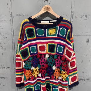 Könnte beinhalten: Ein langärmeliger Pullover mit einem mehrfarbigen Häkeldesign aus quadratischen Motiven und einem durchbrochenen Muster in der Mitte. Das Kleidungsstück zeigt leuchtende Farben wie Rot, Blau, Gelb, Grün und Orange mit Rundhalsausschnitt und Bogenkanten.