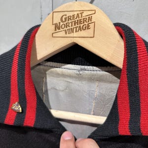 Op de afbeelding: Een zwarte vintage jas met een rood-zwarte gestreepte kraag. De jas hangt aan een houten hanger met de tekst "Great Northern Vintage" erop gegraveerd. Een goudkleurige speld is aan de jas bevestigd.