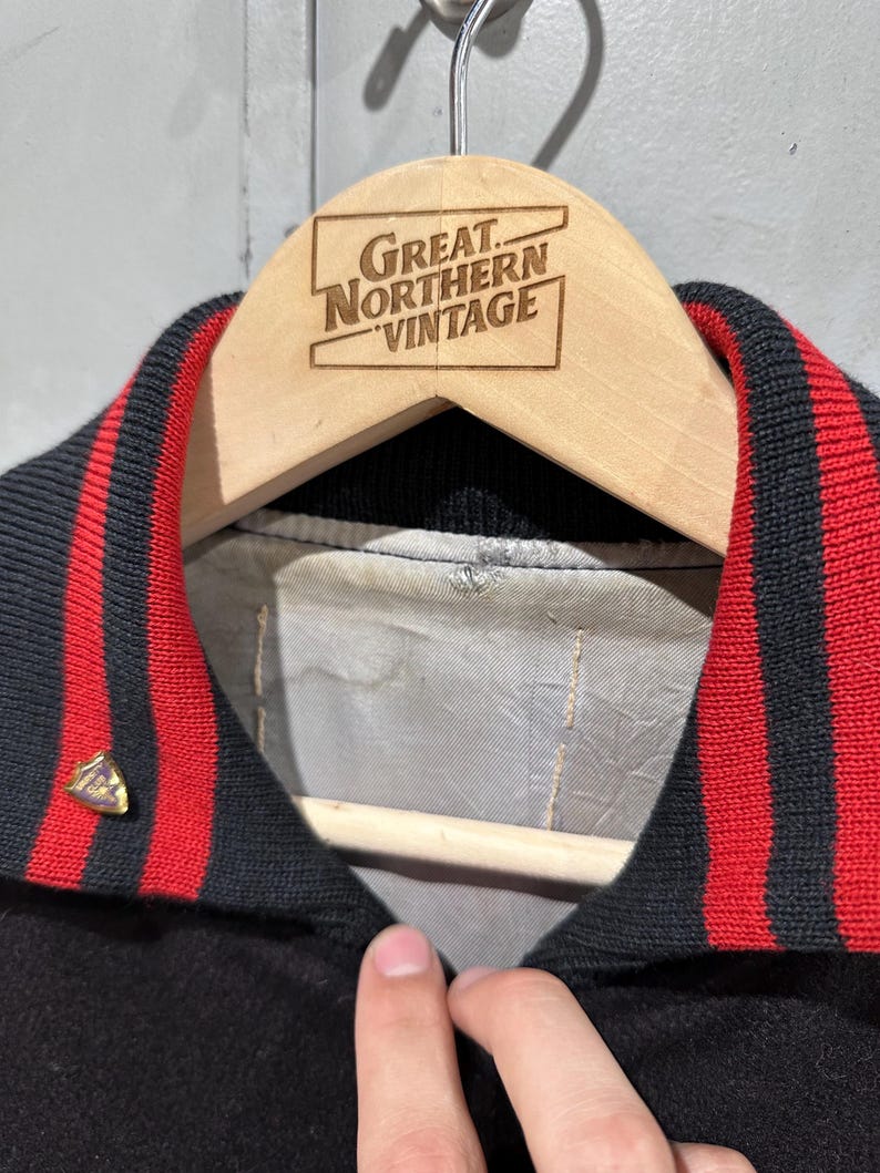 Op de afbeelding: Een zwarte vintage jas met een rood-zwarte gestreepte kraag. De jas hangt aan een houten hanger met de tekst "Great Northern Vintage" erop gegraveerd. Een goudkleurige speld is aan de jas bevestigd.