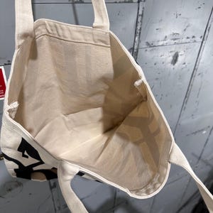 May include: An off-white canvas tote bag with two shoulder straps. The exterior features bold black lettering printed on the fabric. The interior is open and unlined, showing the natural texture of the woven cotton material throughout the bag.