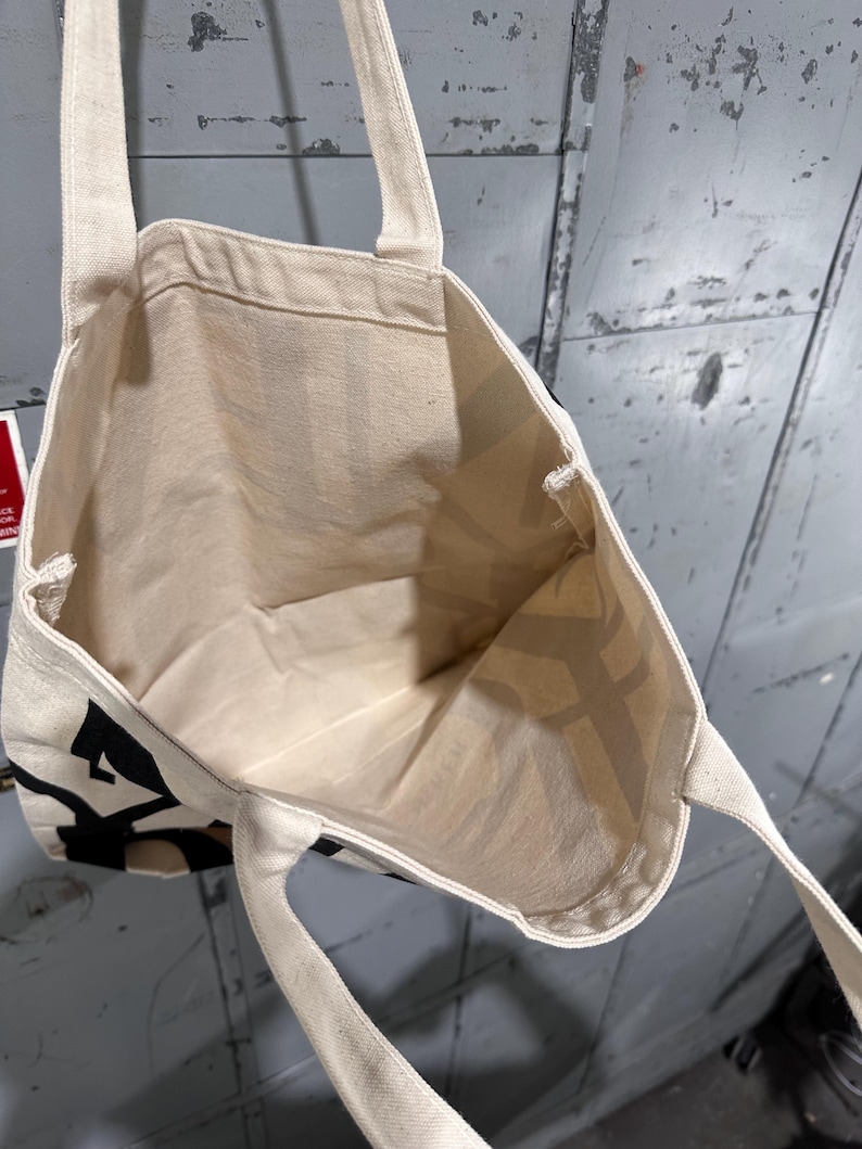 May include: An off-white canvas tote bag with two shoulder straps. The exterior features bold black lettering printed on the fabric. The interior is open and unlined, showing the natural texture of the woven cotton material throughout the bag.
