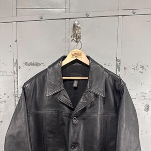 May include: A black leather jacket featuring a classic pointed collar, a button-front closure with visible stitching, and long sleeves. The jacket is displayed on a wooden hanger that has the text Great Northern Vintage printed on it.