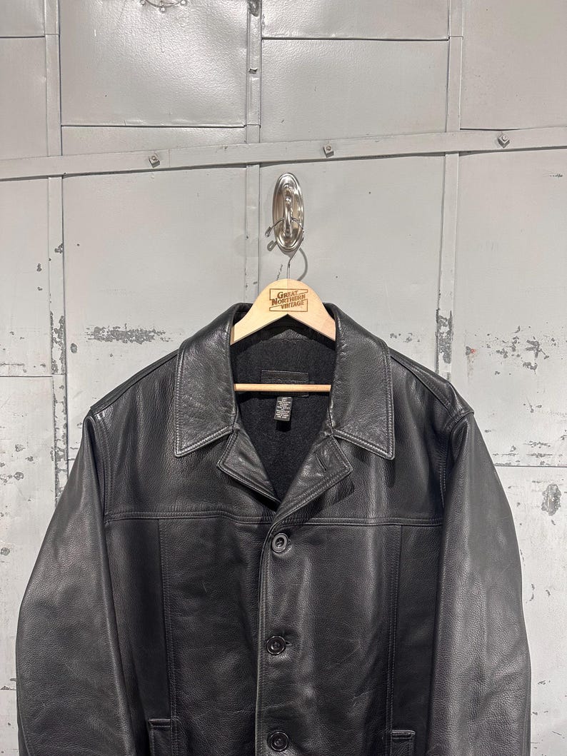 May include: A black leather jacket featuring a classic pointed collar, a button-front closure with visible stitching, and long sleeves. The jacket is displayed on a wooden hanger that has the text Great Northern Vintage printed on it.