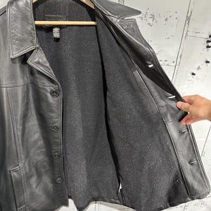 May include: A black leather jacket featuring a pointed collar, a button-front closure, and long sleeves. The interior is lined with a dark gray felt-like material. The jacket includes two side pockets and a visible brand tag at the inner neckline.
