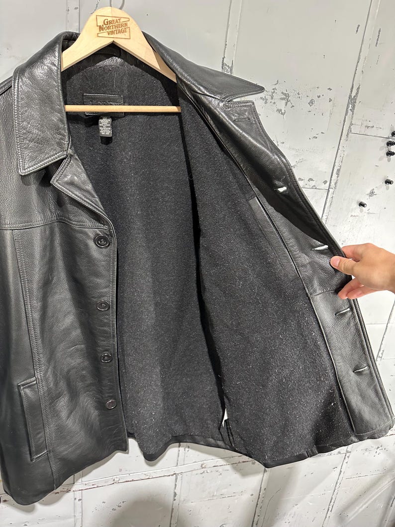May include: A black leather jacket featuring a pointed collar, a button-front closure, and long sleeves. The interior is lined with a dark gray felt-like material. The jacket includes two side pockets and a visible brand tag at the inner neckline.